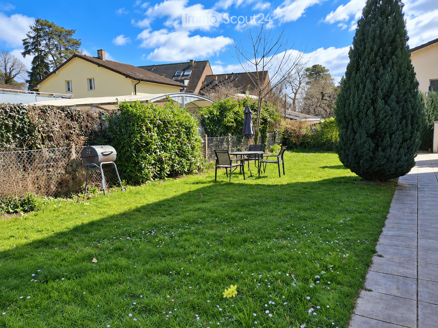 Well-maintained garden with a grassy lawn, a patio area with a table and chairs, a grill, and surrounding hedges and trees.