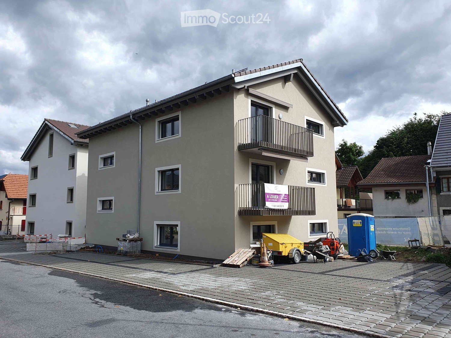Two-story house with grey facade, multiple windows, and a balcony on the upper floor. The building is surrounded by a paved area with construction materials and equipment.