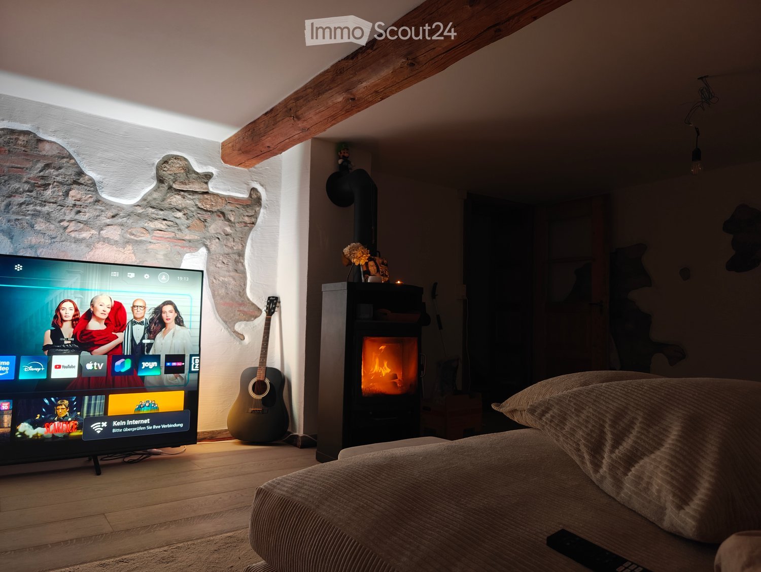 Cozy living room with exposed wooden beams, a stone wall, a fireplace, a television, a guitar, and a sofa.