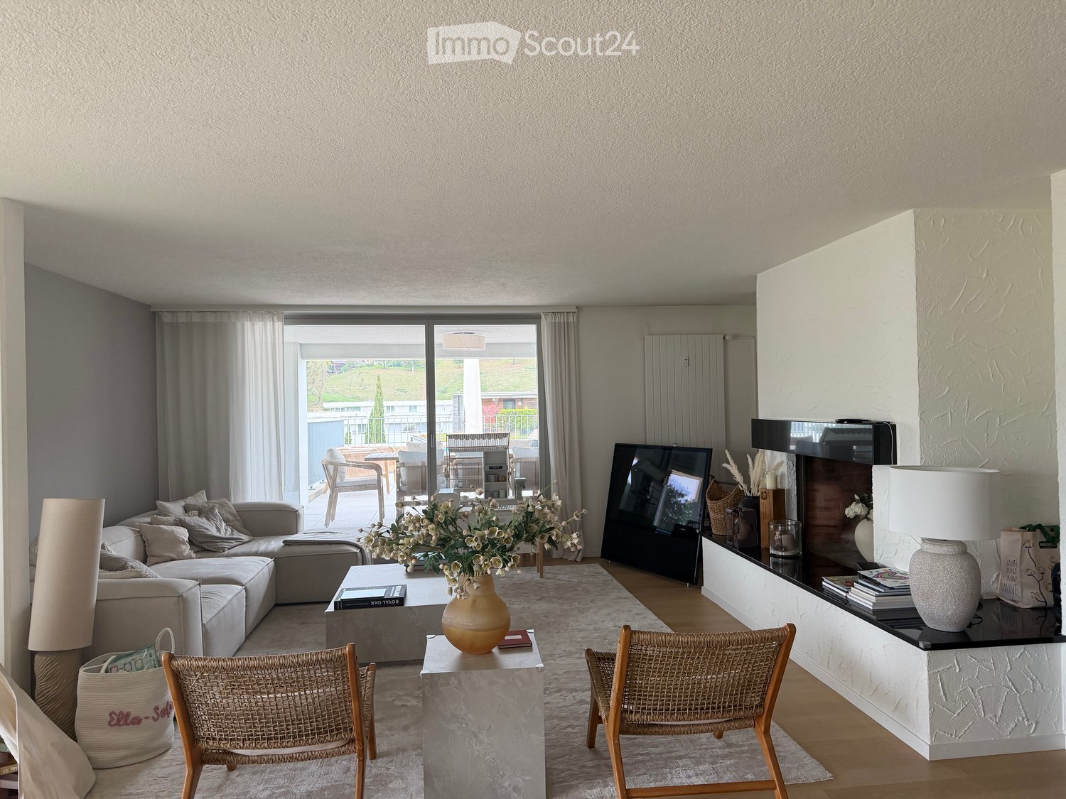 Modern living room with a grey sectional sofa, coffee table, wicker chairs, a large TV, and a fireplace with decorative items. Large windows offer a view of the outdoor area.