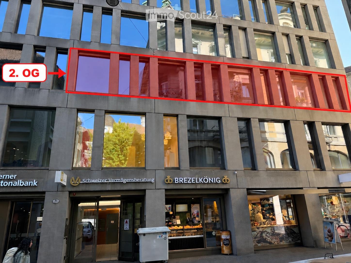 Modern multi-story building with large windows and signage for 'Schweizer Vermögensberatung' and 'BREZELKÖNIG'. The ground floor features a bakery with visible pastries in the display window.