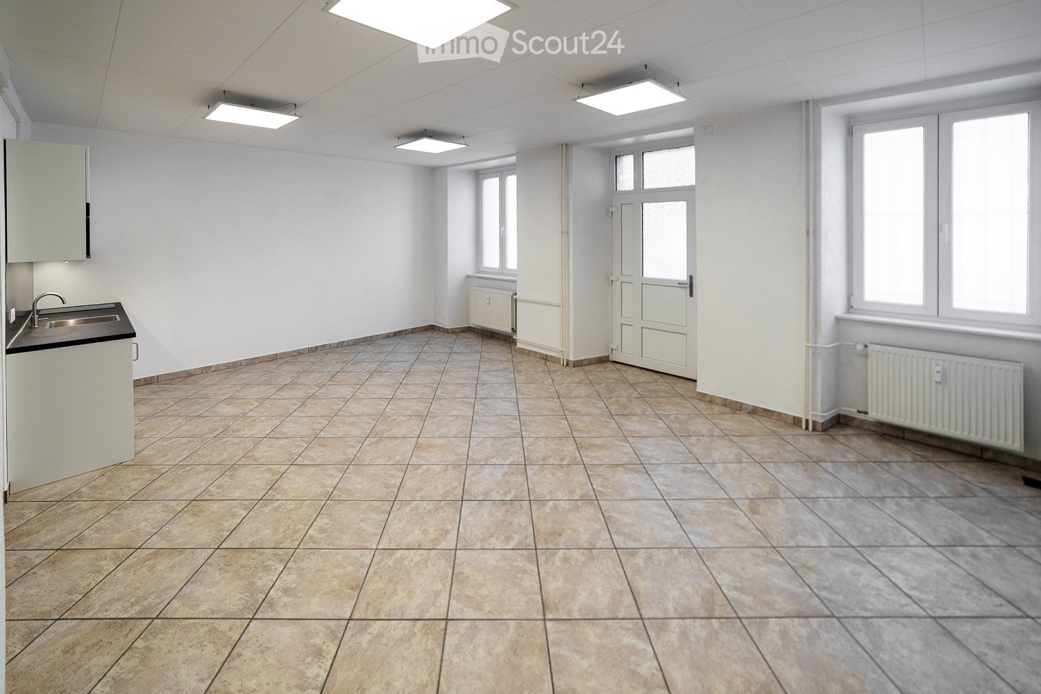 Empty room with tiled floor, white walls, and a small sink area. The room has several windows and a door leading to another space.