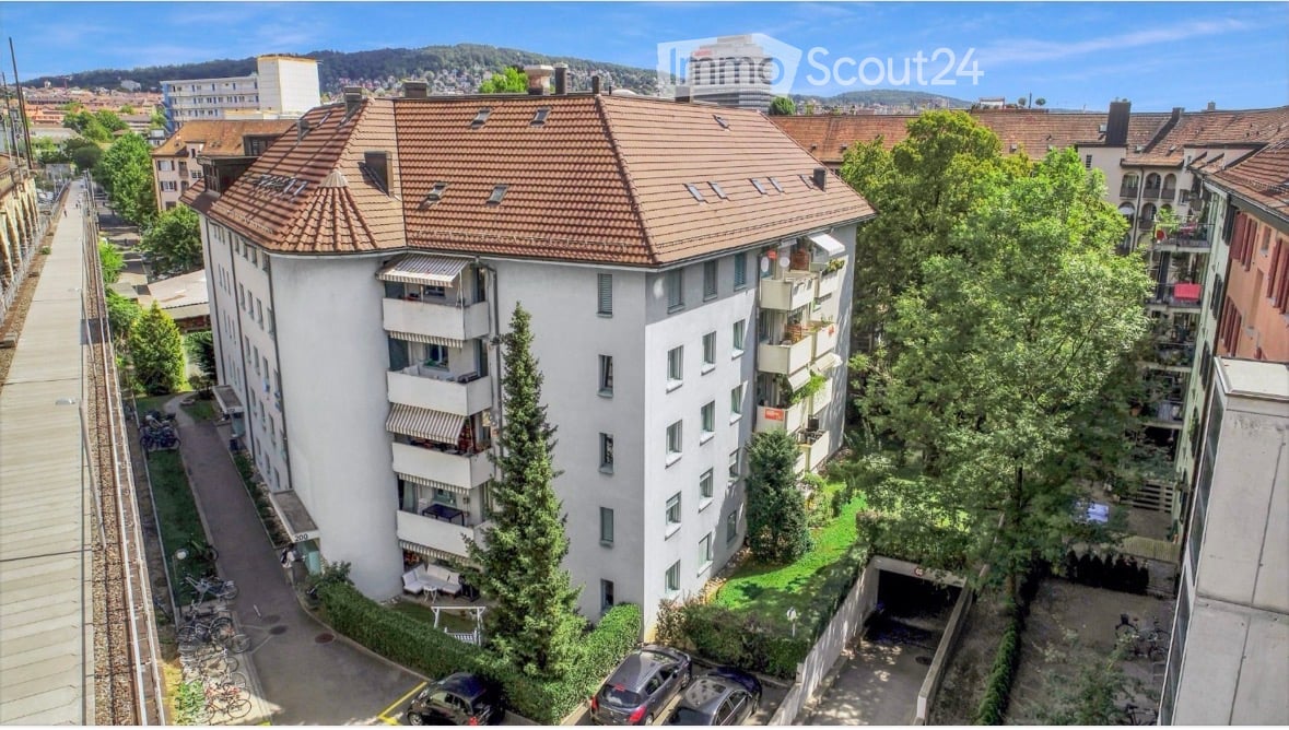 Multi-story residential building with a red-tiled roof, balconies, and surrounding greenery. Includes a parking area and garage.