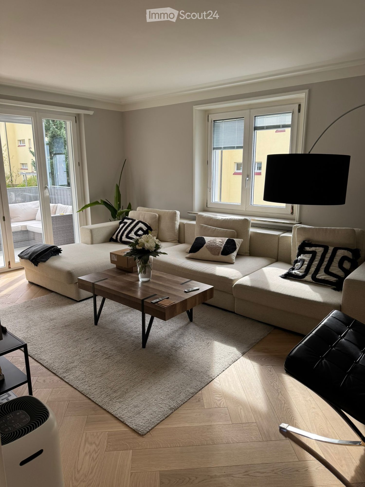 Modern living room with a large sectional sofa, coffee table, floor lamp, and a black leather chair. The room features large windows and a sliding glass door leading to a balcony.