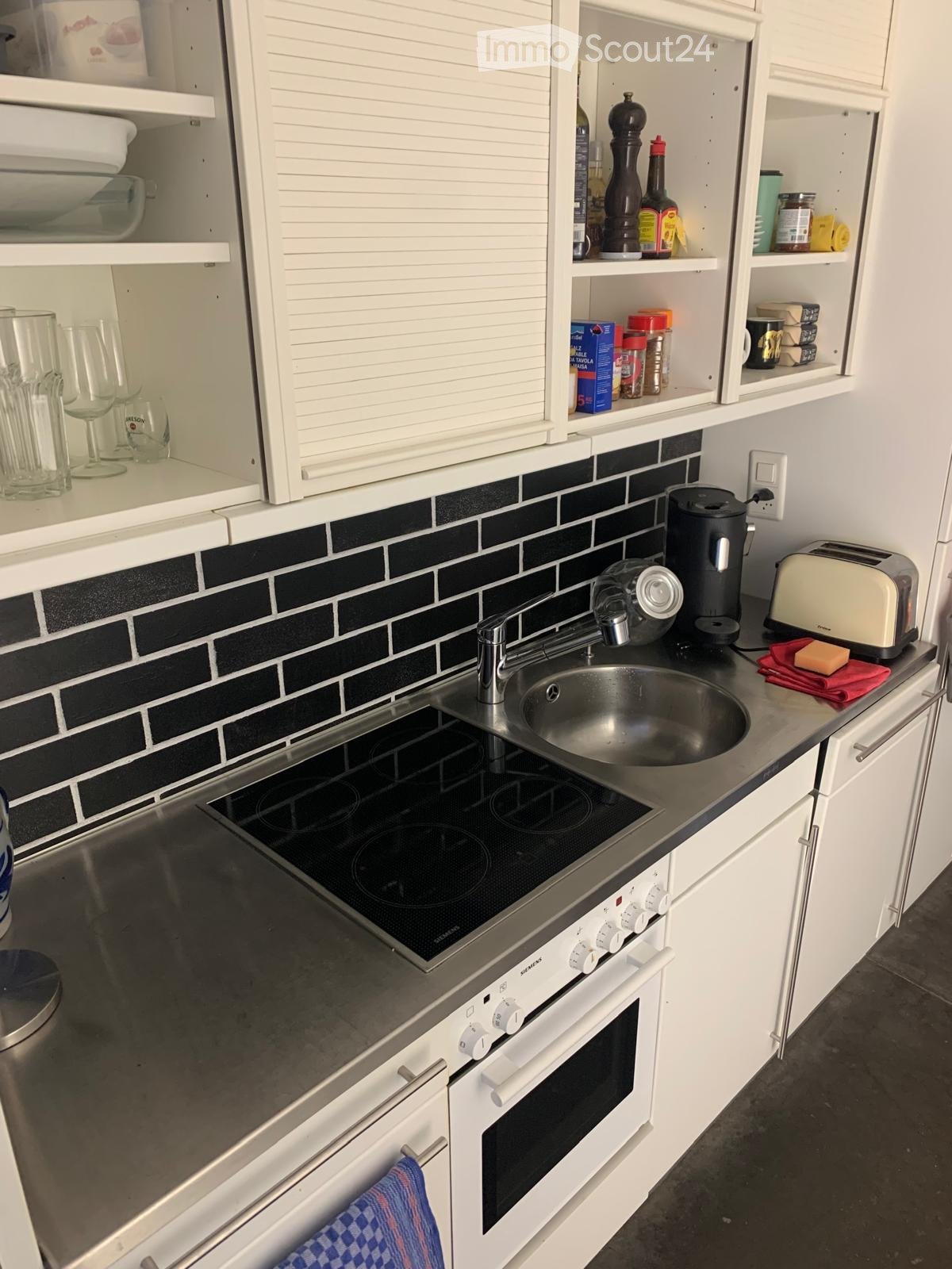Modern kitchen with white cabinets, stainless steel sink, induction cooktop, oven, coffee maker, and toaster. Shelves hold various kitchen items including bottles, cups, and food containers.