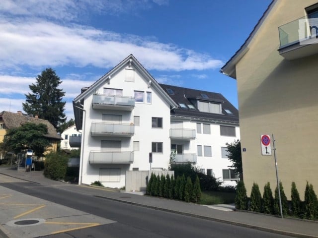 Modern multi-story apartment building with white exterior, multiple balconies, and surrounding greenery.