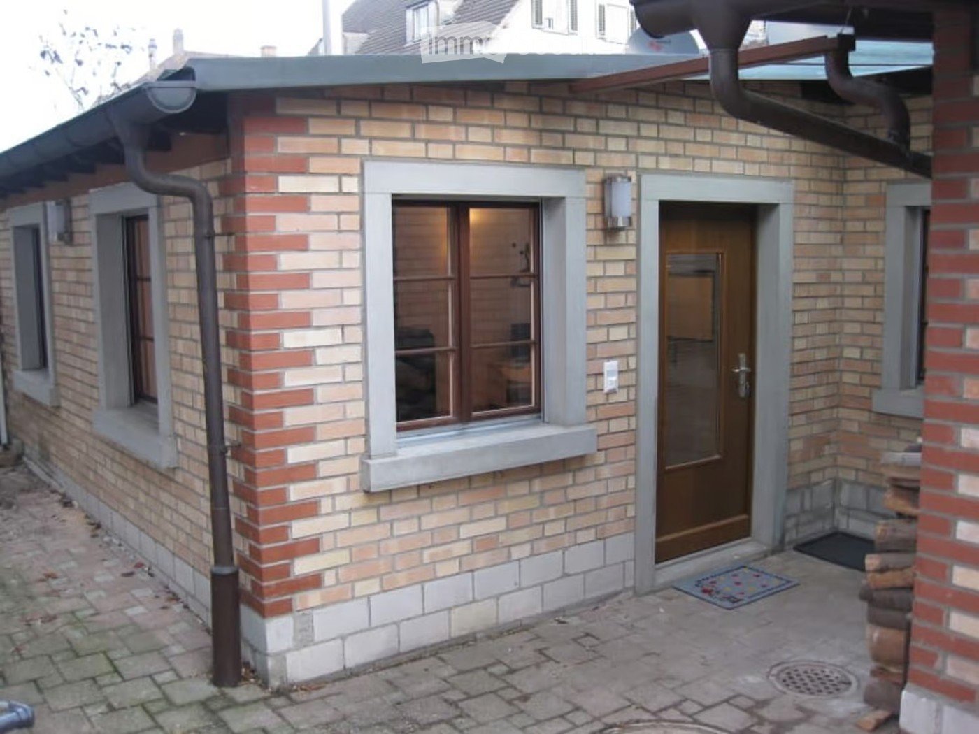 Exterior view of a small brick building with a flat roof, featuring a wooden door, a window with a view into a room, and a paved stone pathway.