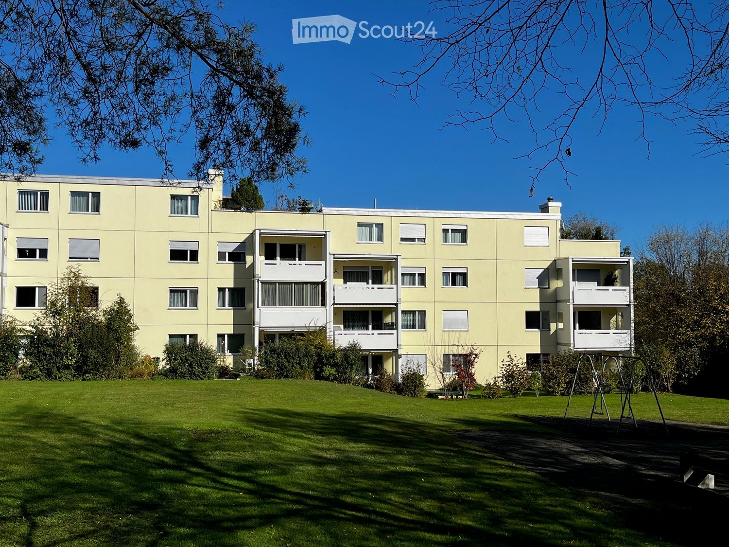 A multi-story residential building with light yellow walls, multiple windows, and balconies, surrounded by a well-maintained lawn and trees.