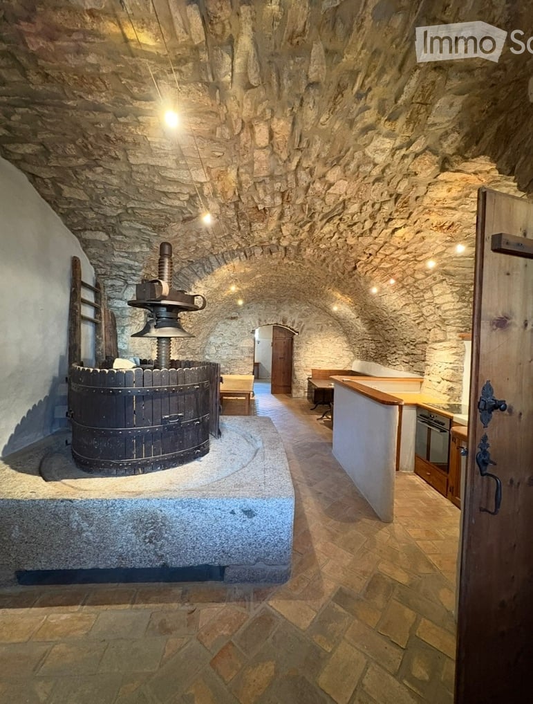 Stone-walled room with a large wooden wine press, a counter, a stool, and an oven.