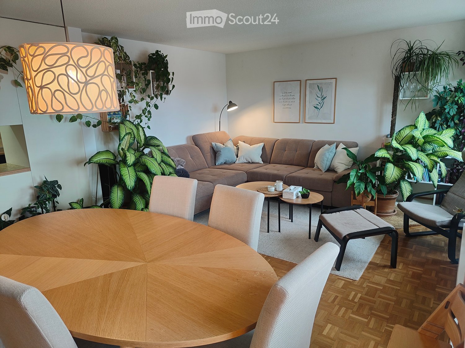 A modern living room with a large sectional sofa, coffee table, dining table with chairs, and several potted plants. The room features wooden flooring and a hanging lamp with a unique design.