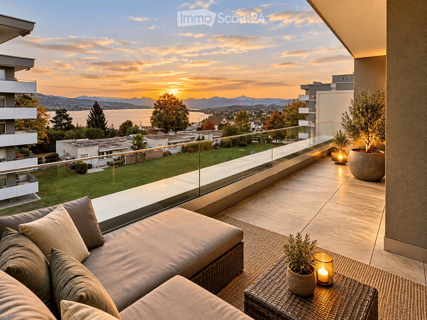 Modern balcony with a view of a lake and mountains at sunset, featuring a wicker sofa, a small table with a candle, and potted plants.