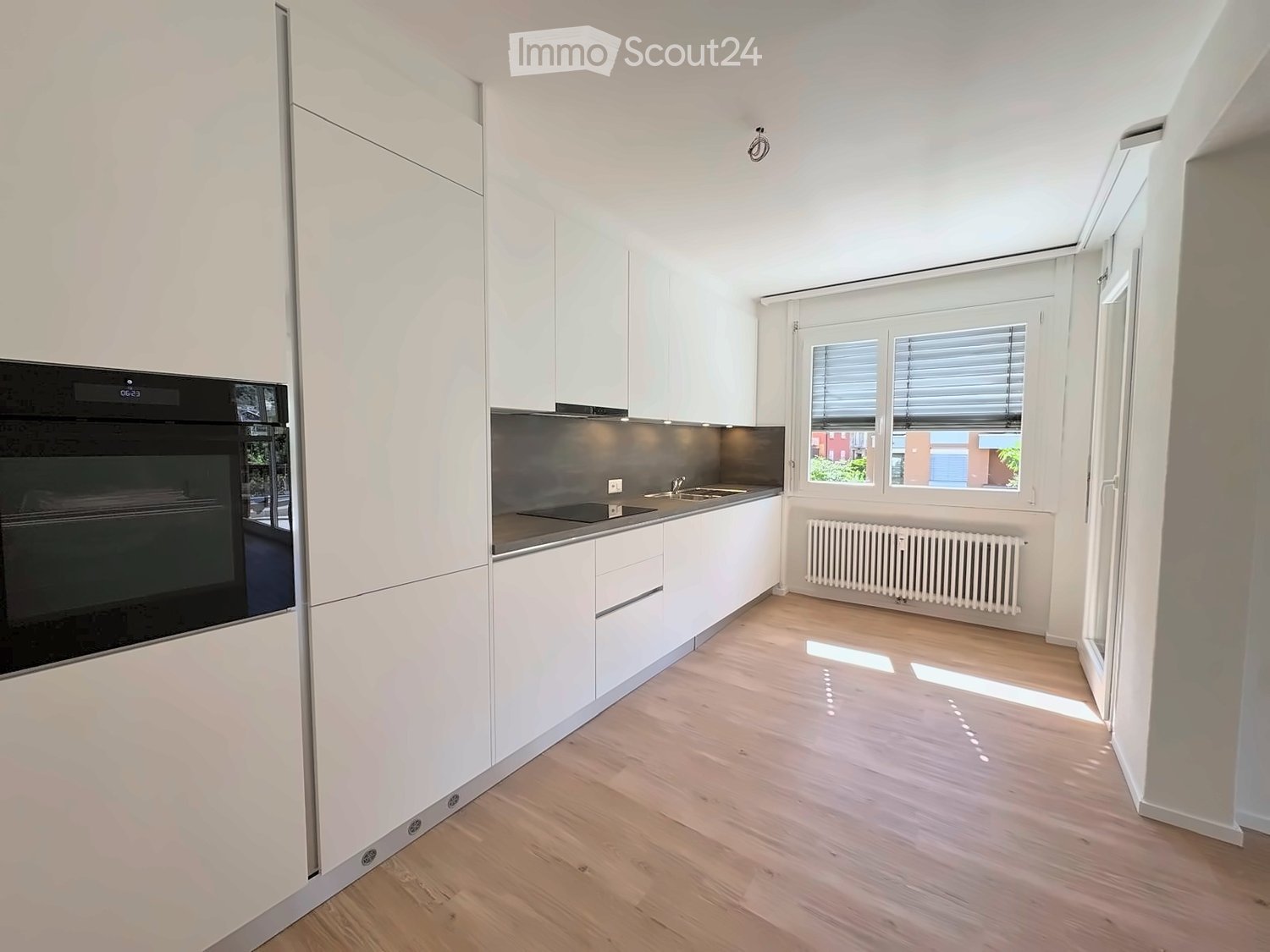 Modern kitchen with white cabinets, wooden flooring, and a black oven. Features a sink, radiator, and large windows with blinds.