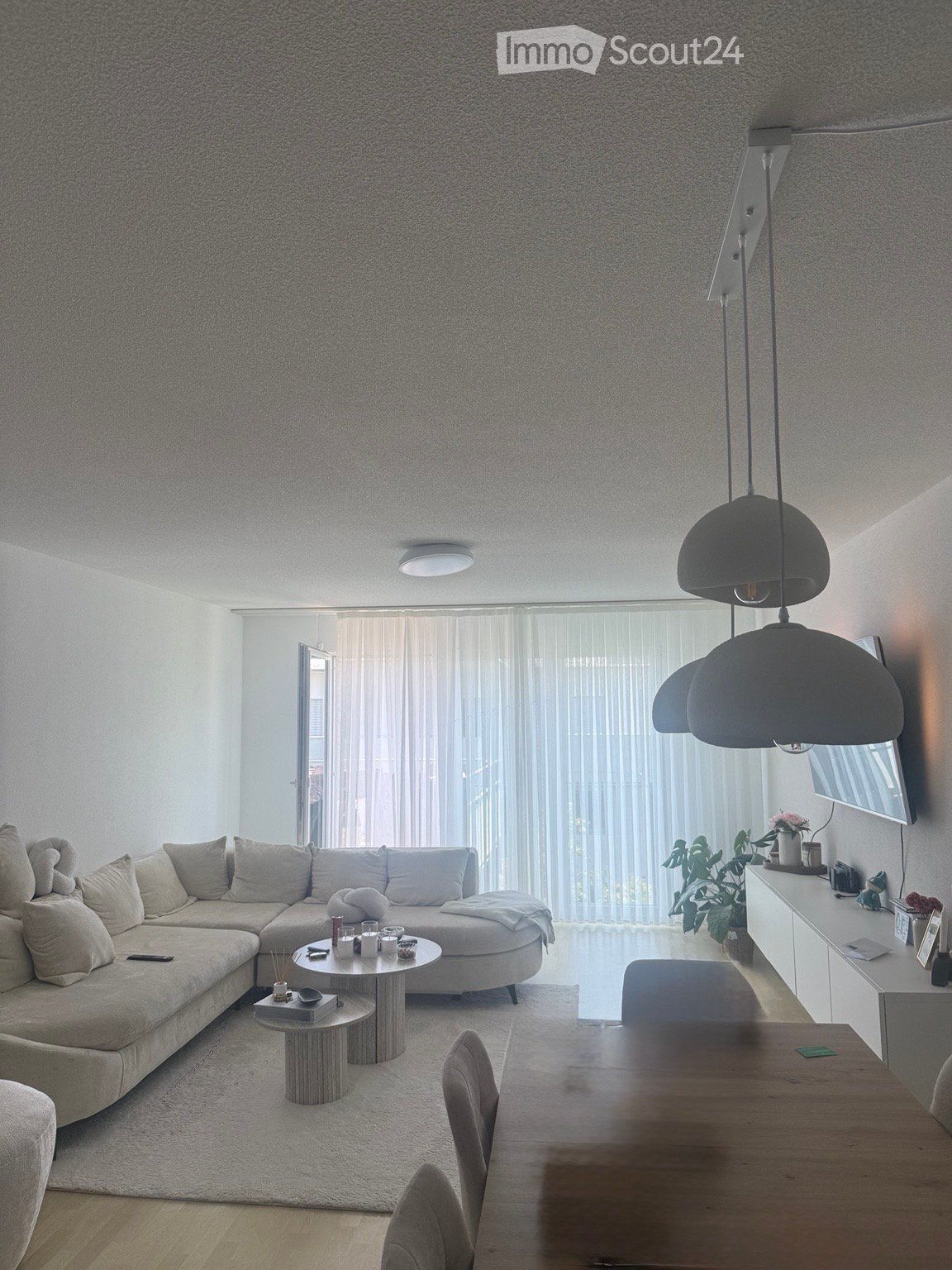 Modern living room with a large sectional sofa, coffee table, dining table, pendant lights, and a TV screen. The space features large windows with sheer curtains and a neutral color palette.