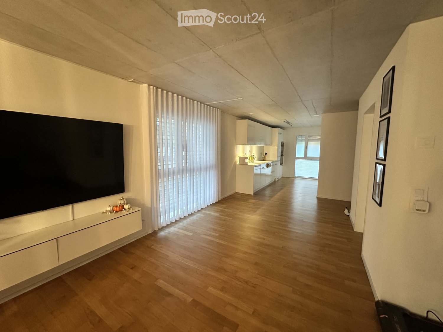 Open-plan living and kitchen area with wooden flooring, white kitchen cabinets, a large television, and framed pictures on the wall.
