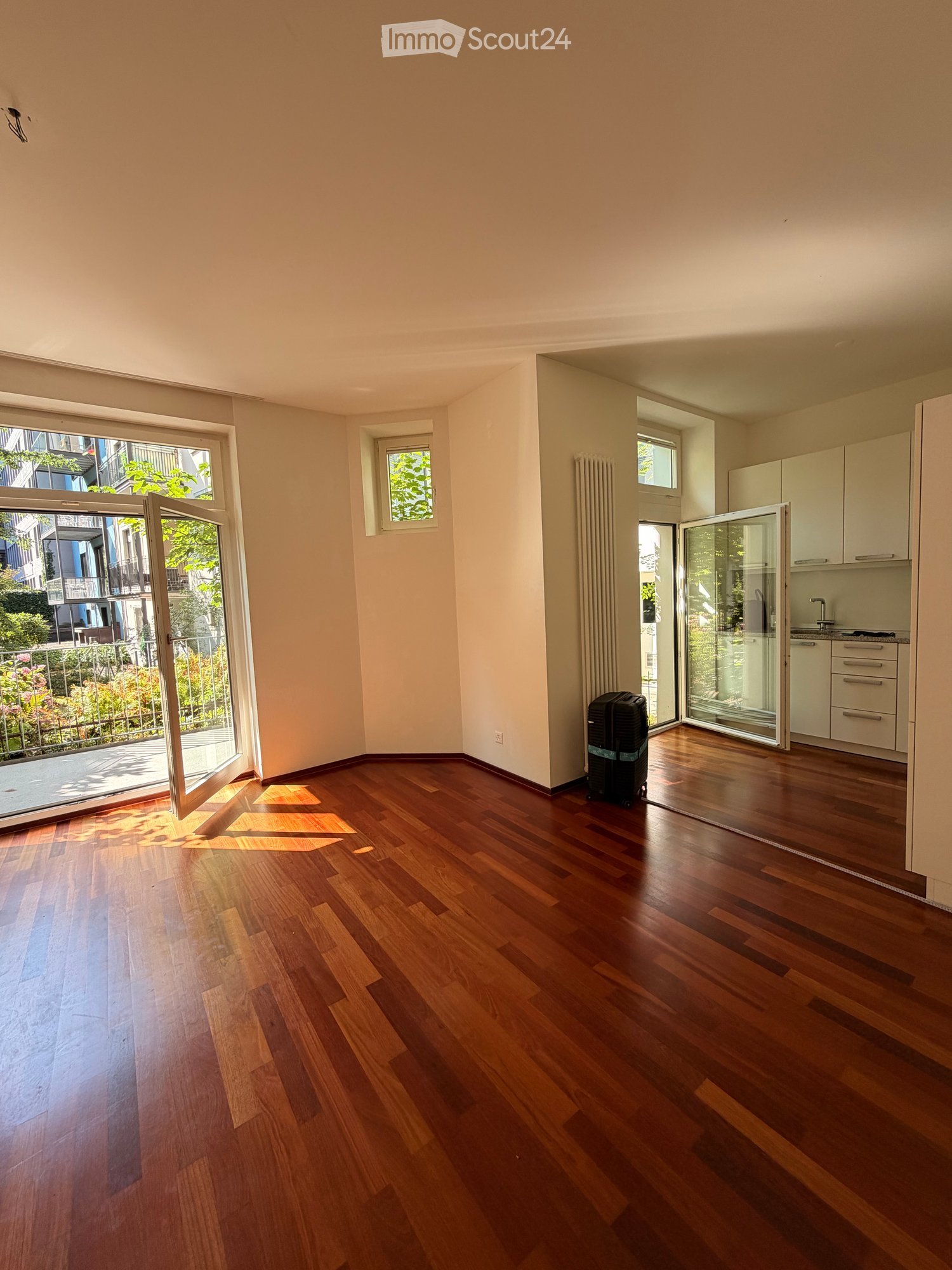 Empty room with wooden flooring, large windows, and a view of a balcony area with greenery.