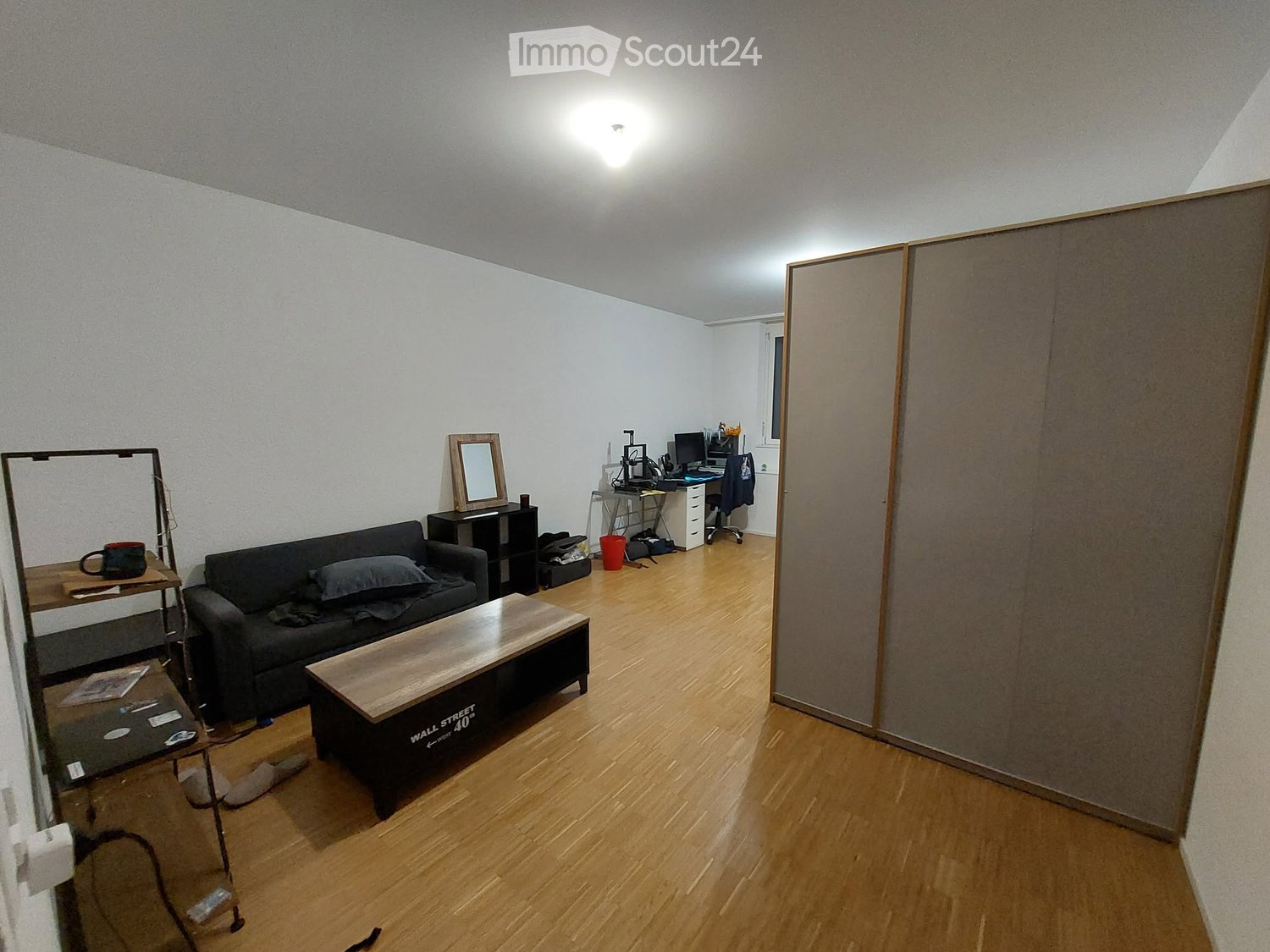 A modern, well-lit living room with wooden flooring, a grey sofa, a wooden coffee table, a desk with a laptop, and a mirror on a side table.