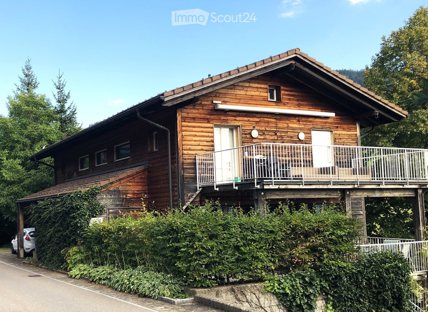 Two-story wooden house with a balcony, surrounded by greenery and trees.