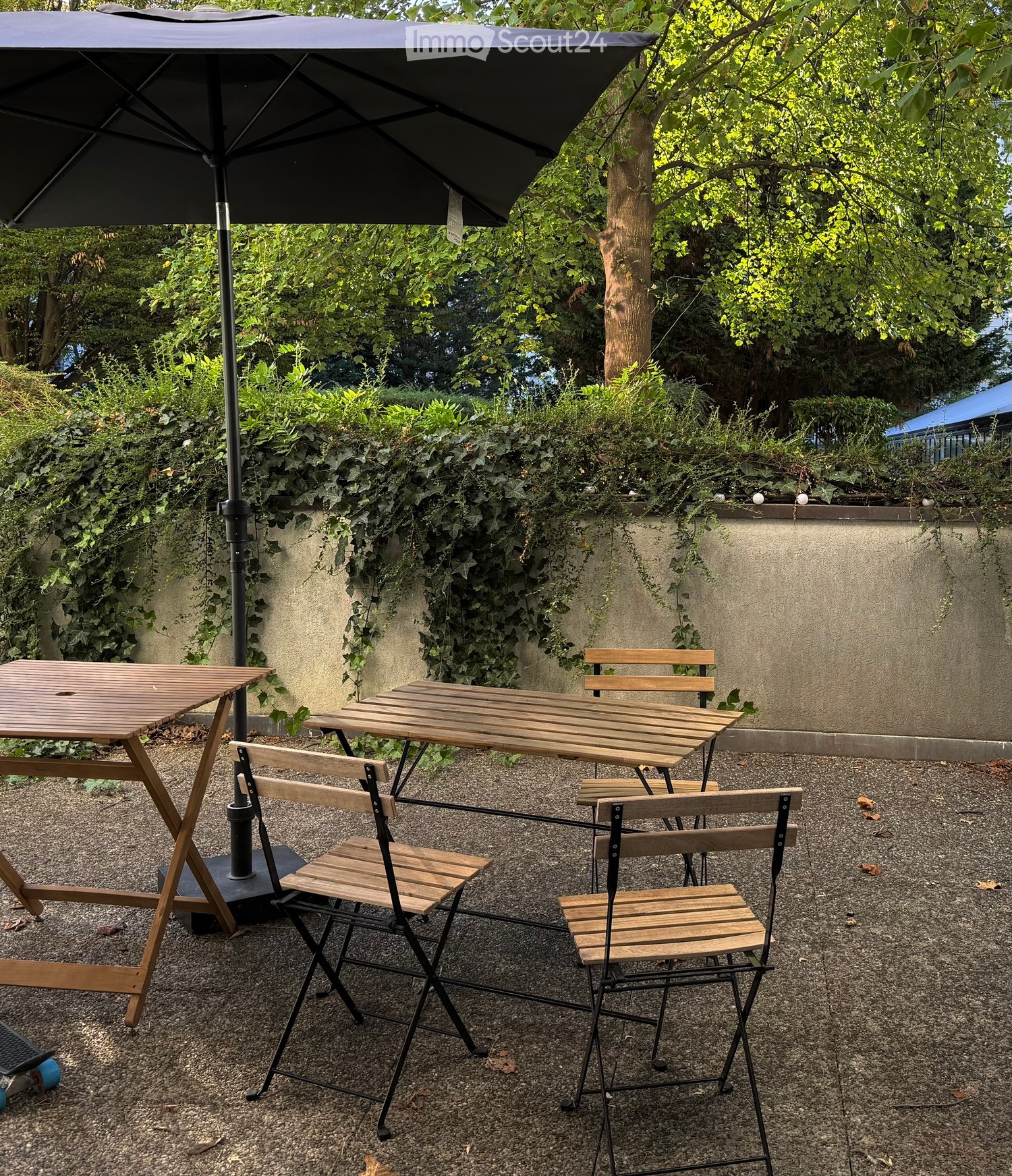 Garden area with gravel ground, two wooden tables, four wooden chairs, and a large black umbrella providing shade.
