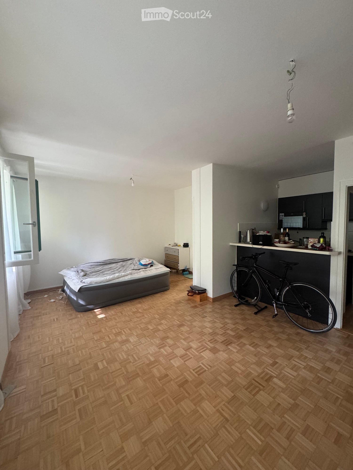 A compact studio apartment with a wooden herringbone floor, a bed with a grey duvet, a small kitchen area with a counter, microwave, fridge, and a bicycle leaning against the wall.