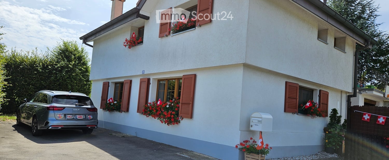 Two-story white house with red shutters, flower boxes, and a parked car in front.
