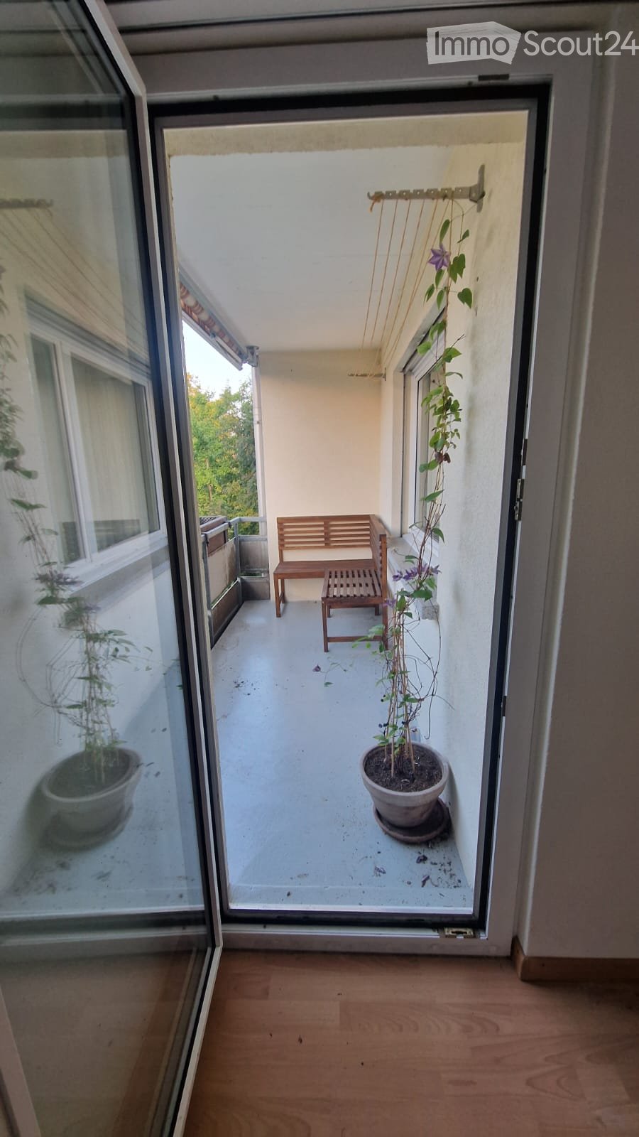 Balcony with a wooden bench, small table, and potted plants, overlooking a green area.