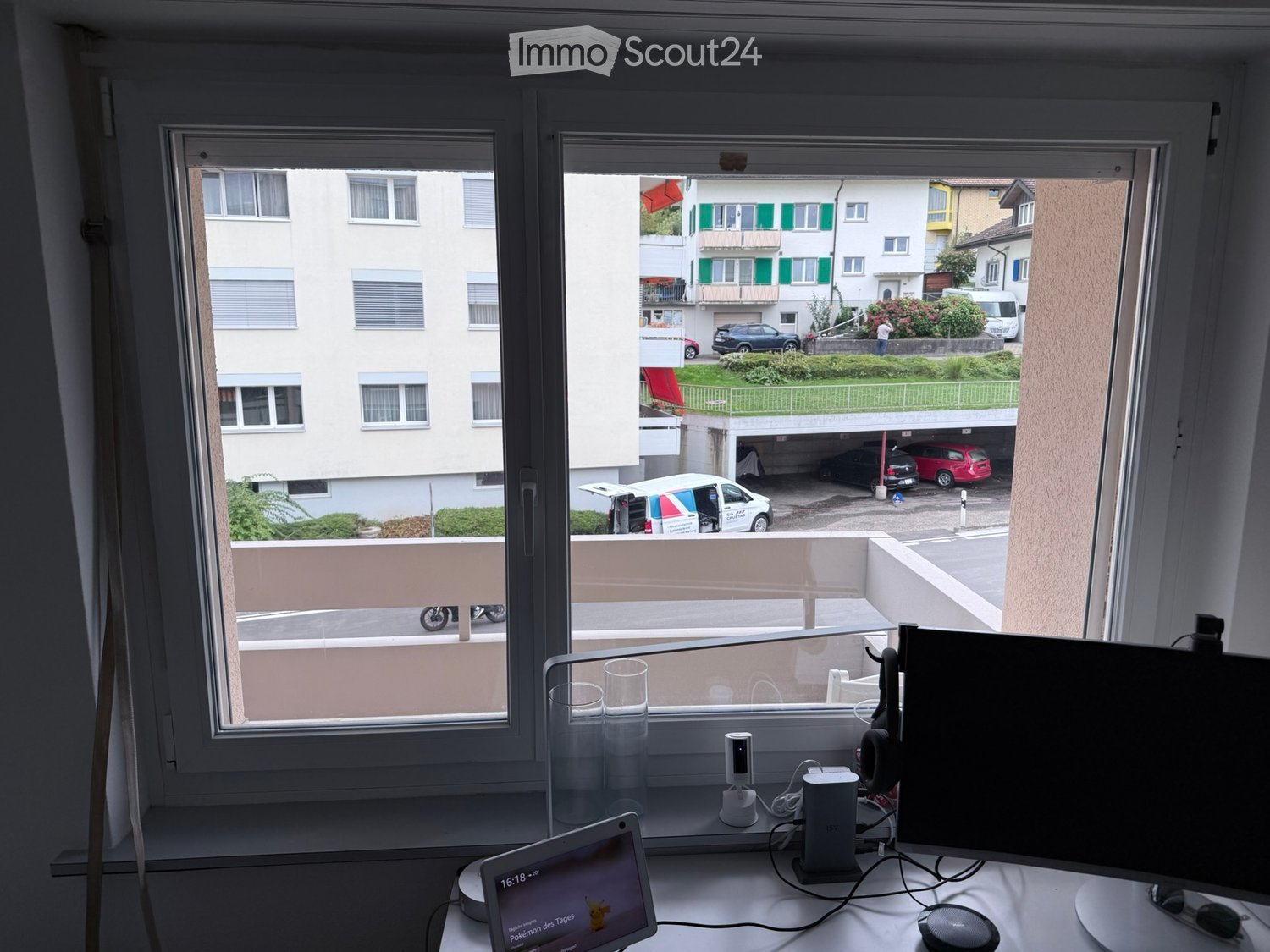 A modern workspace with a laptop, screen, headphones, and laptop stand on a white desk, overlooking a balcony with a view of a residential area and parked cars.