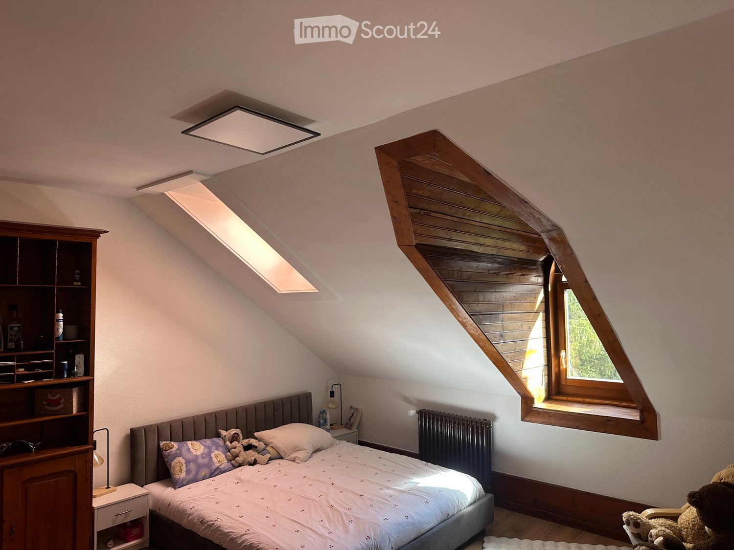 Cozy attic bedroom with slanted wooden ceiling, skylight windows, grey upholstered bed, nightstand with lamp, bookshelf, and stuffed animals on the bed and floor.