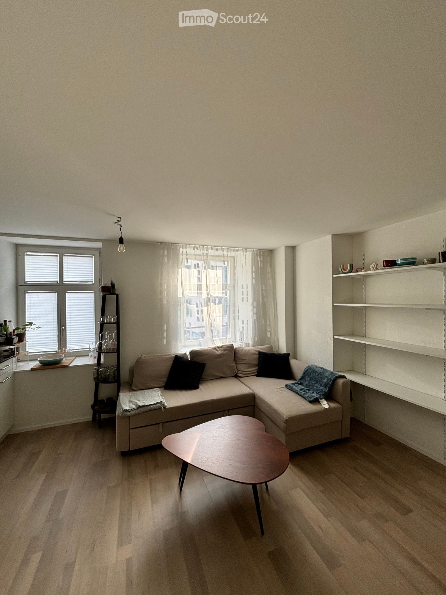 Modern living room with a beige sectional sofa, wooden coffee table, shelves, and large windows with curtains.