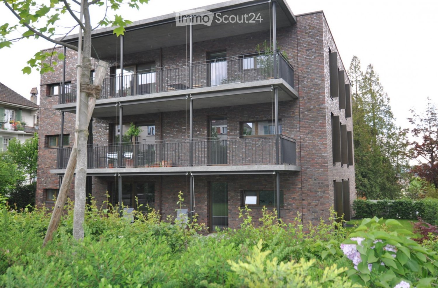 Modern multi-story brick building with balconies and large windows, surrounded by greenery.