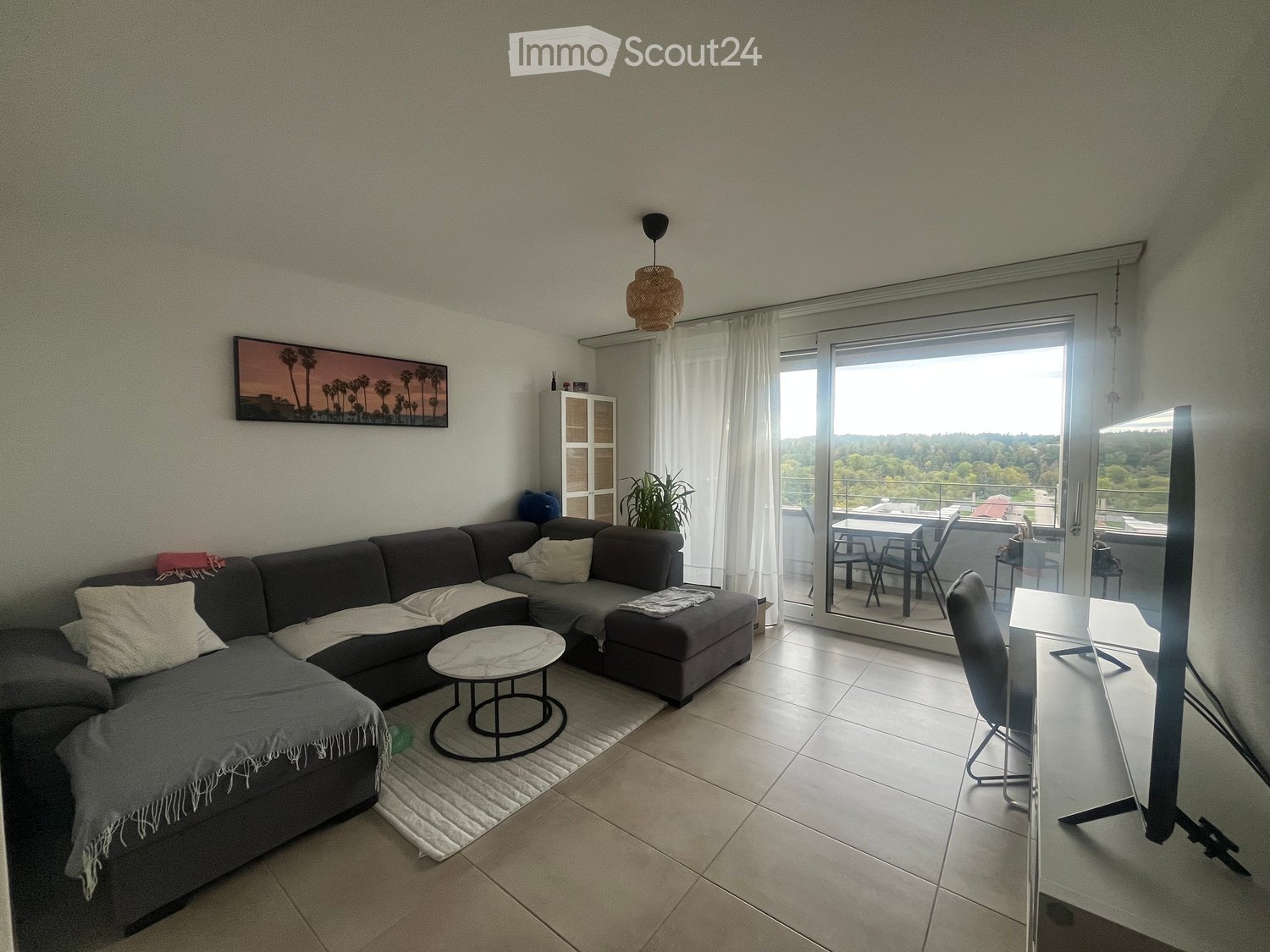 Modern living room with a grey sectional sofa, coffee table, TV screen, and a view of greenery through large windows and a sliding glass door leading to a balcony.