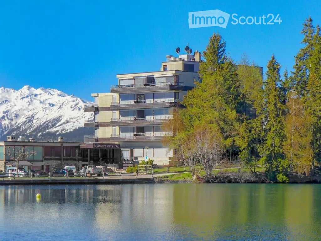 Modern multi-story building with balconies overlooking a lake, surrounded by trees and mountains.