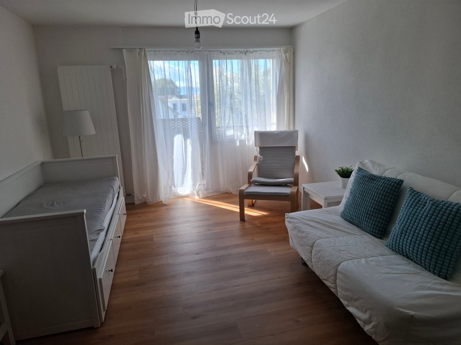 A cozy living room with a sofa, a chair, a small table, a lamp, a bed, and a potted plant. The room features wooden flooring and large windows with white curtains.