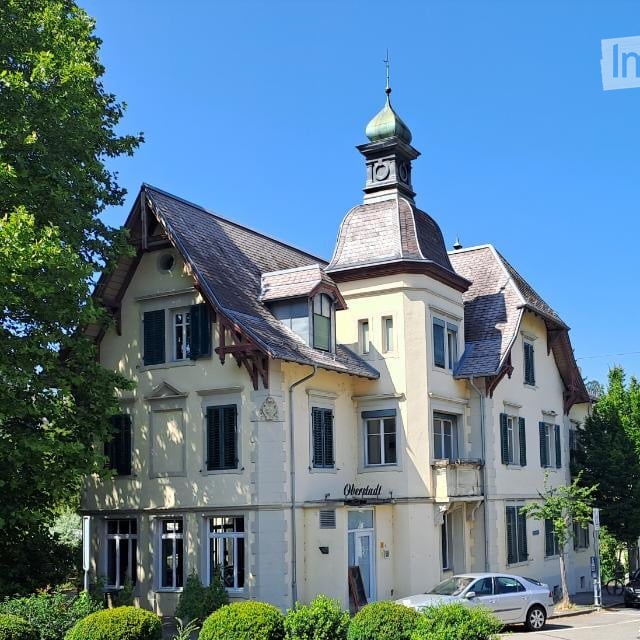A two-story building with a distinctive dome-shaped roof and clock tower, surrounded by greenery and a parked car.