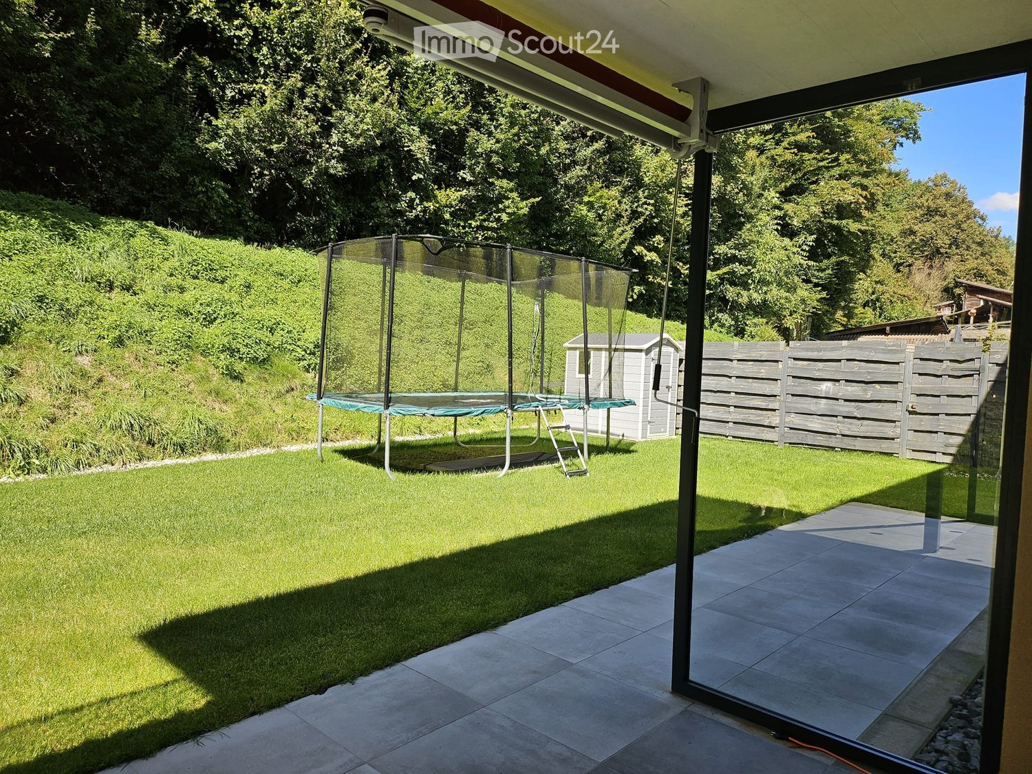 Garden with a trampoline, wooden shed, and wooden fence, surrounded by lush greenery and trees.