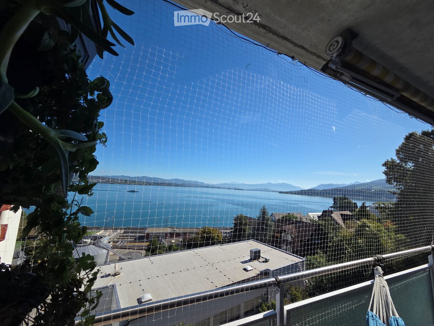 Balcony with a net screen, overlooking a body of water and distant mountains, with a railing and some greenery visible.