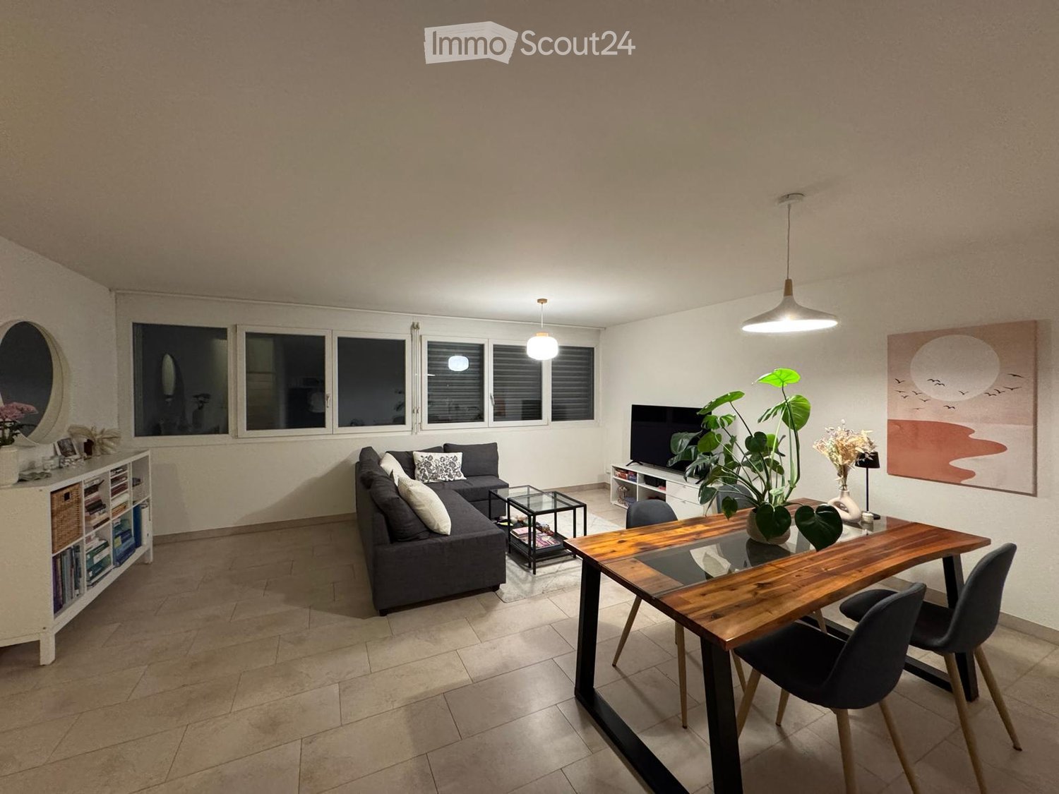 Modern living and dining area with a grey sectional sofa, wooden dining table, glass coffee table, TV, and decorative plants.