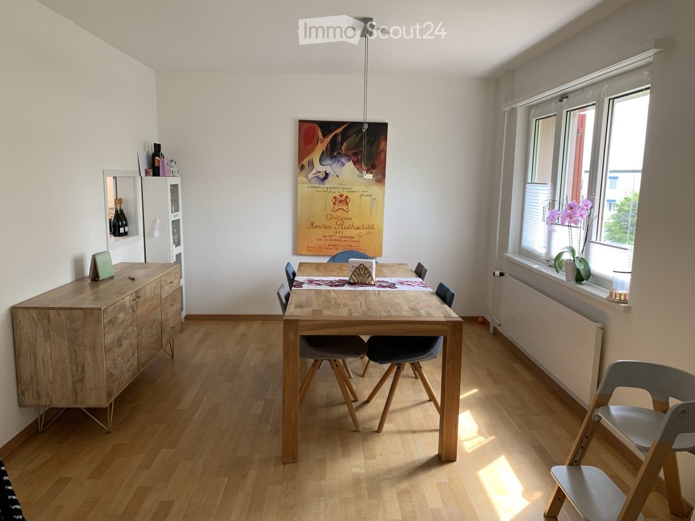 Bright dining area with wooden floor, large window, dining table with chairs, cabinet, and decorative items.