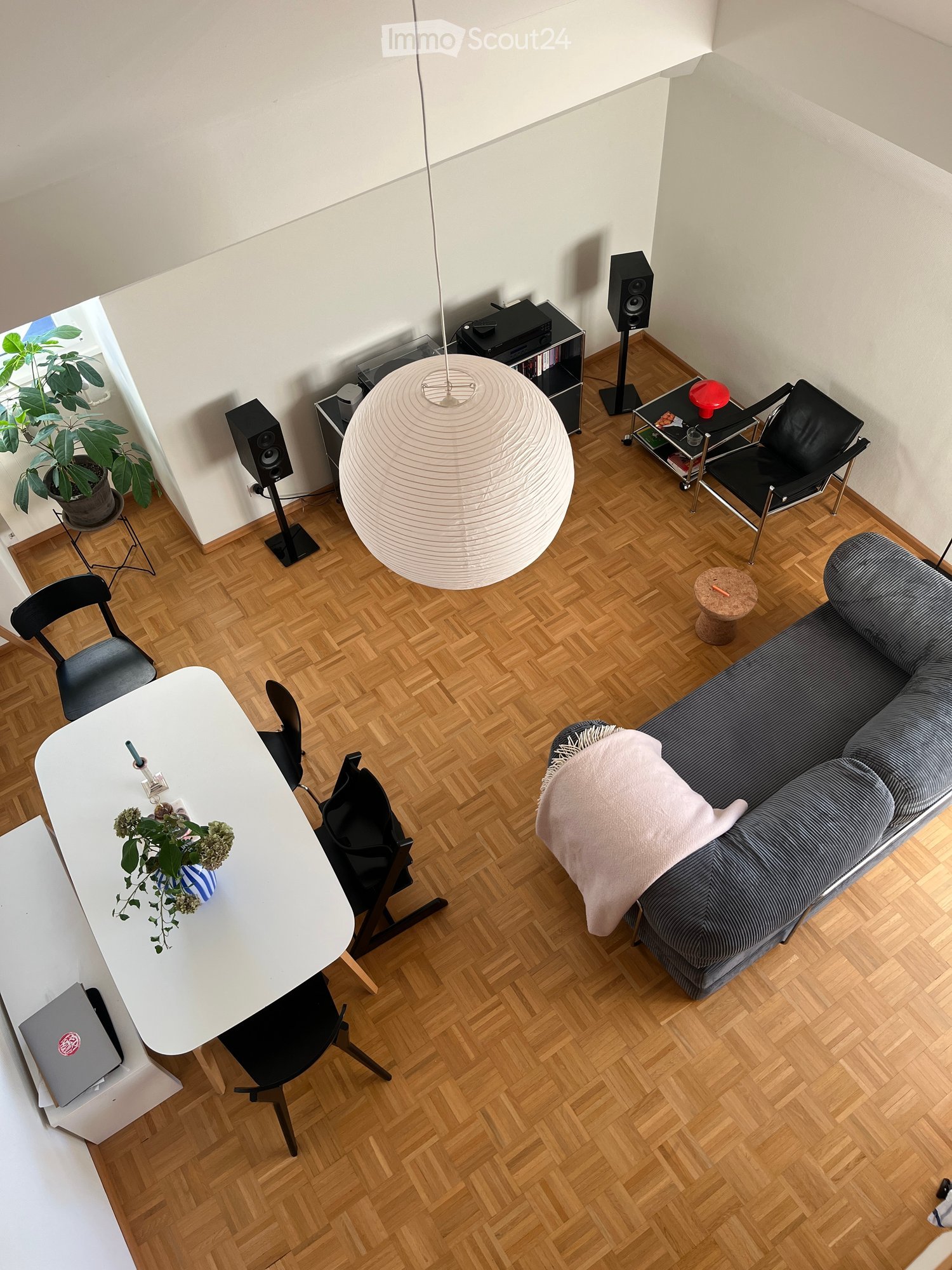 A modern living room with a grey sofa, white coffee table, black chairs, a laptop, a large hanging lamp, speakers, and a plant in the corner.
