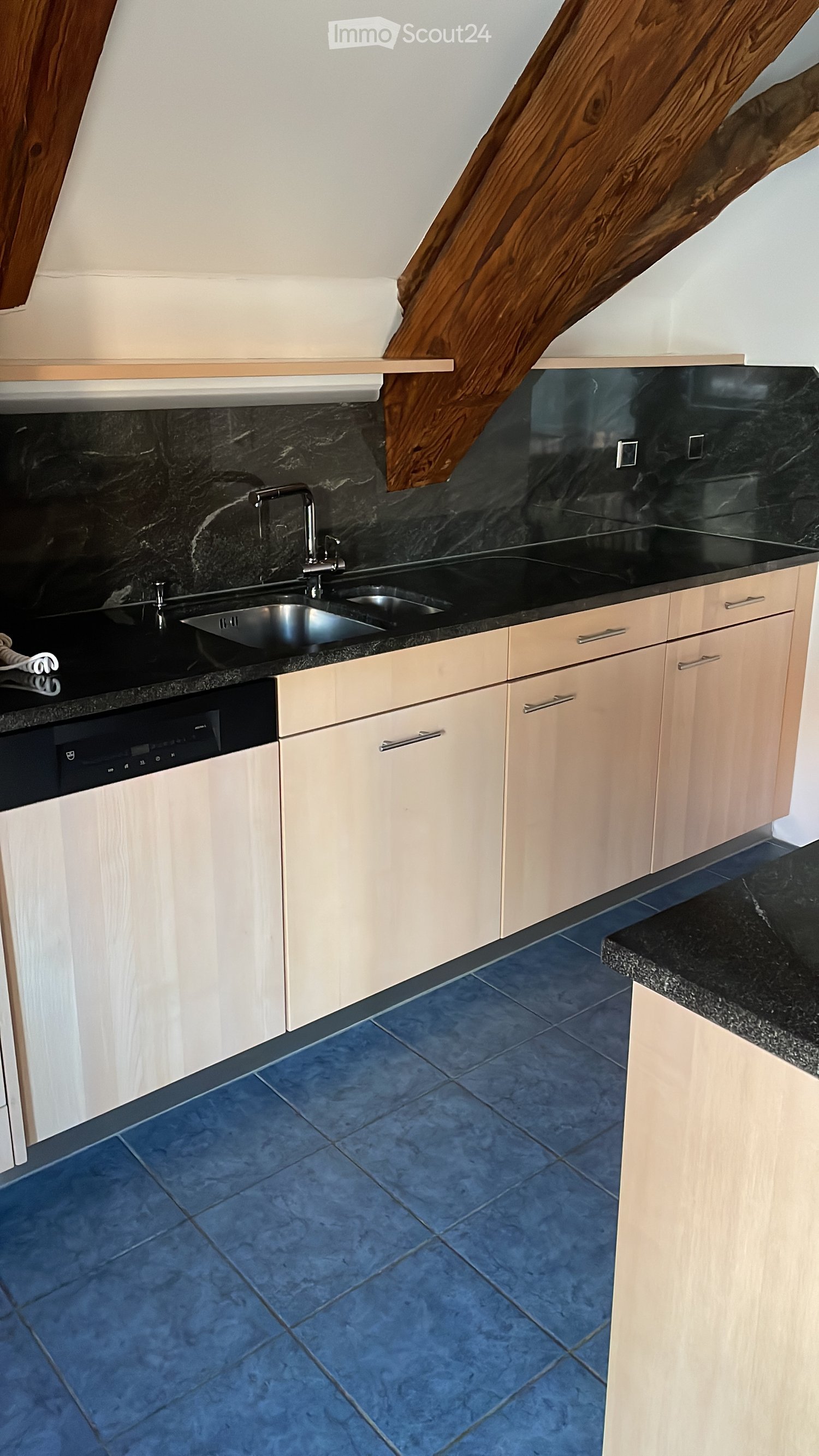 Modern kitchen with light wood cabinets, black granite countertops, stainless steel sink, and dishwasher. Blue tiled floor.