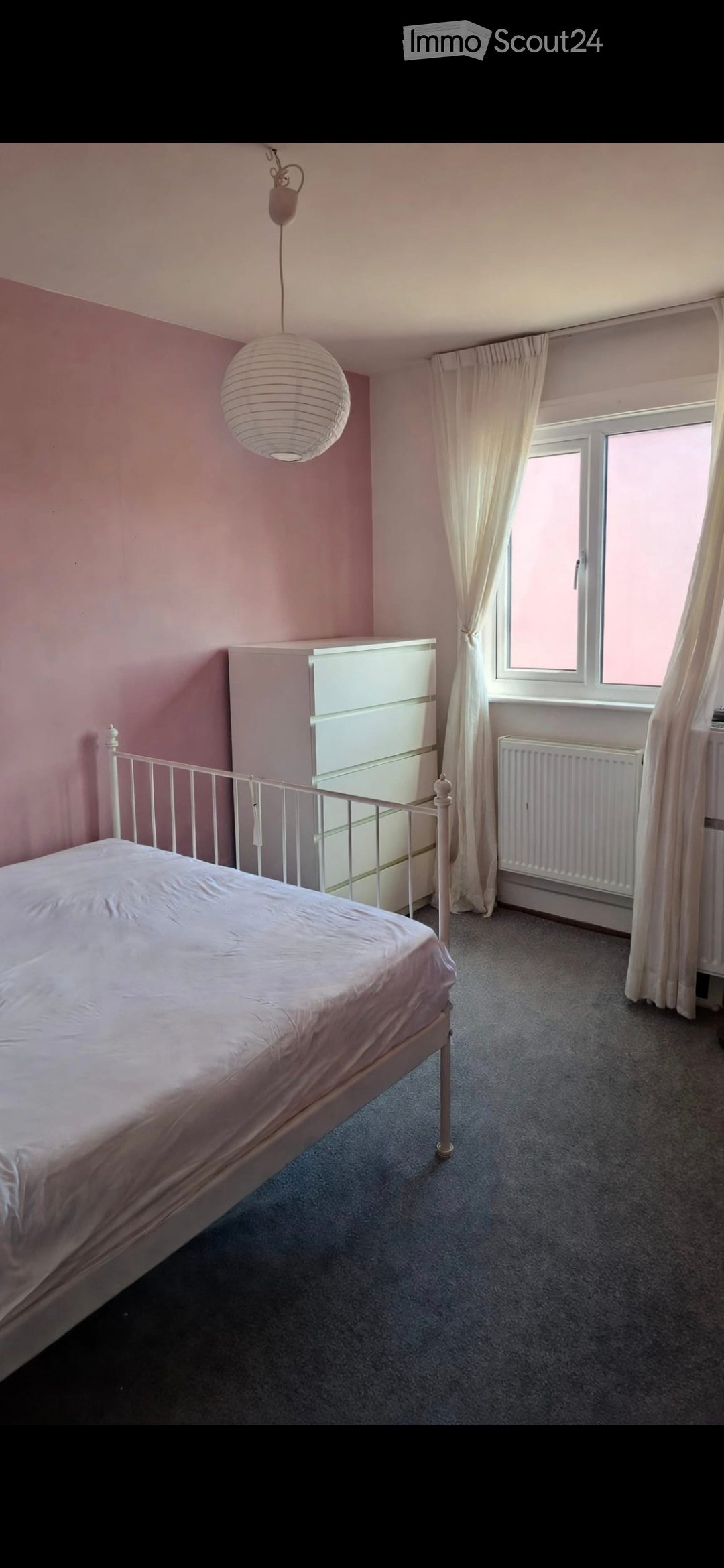 A furnished bedroom with a single bed, white wardrobe, radiator, and a hanging paper lantern light fixture. The room features light pink walls, grey carpet, and white curtains on the window.