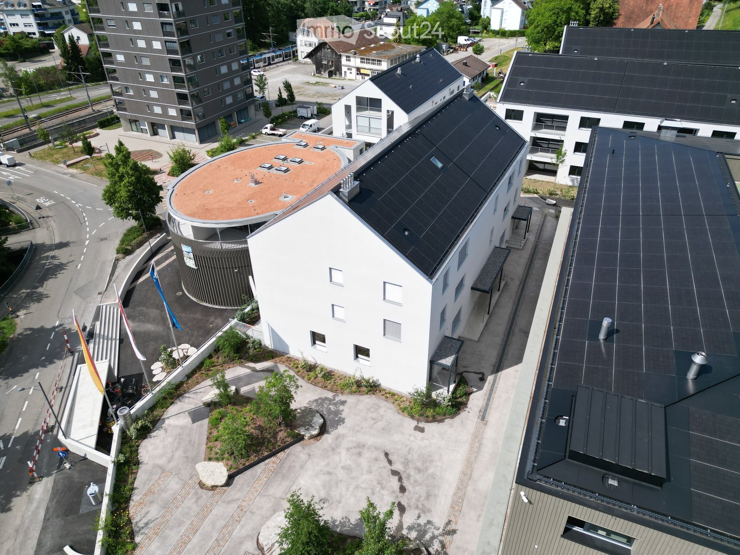 Aerial view of a modern multi-story building complex with solar panels on the roof, surrounded by other structures and greenery.