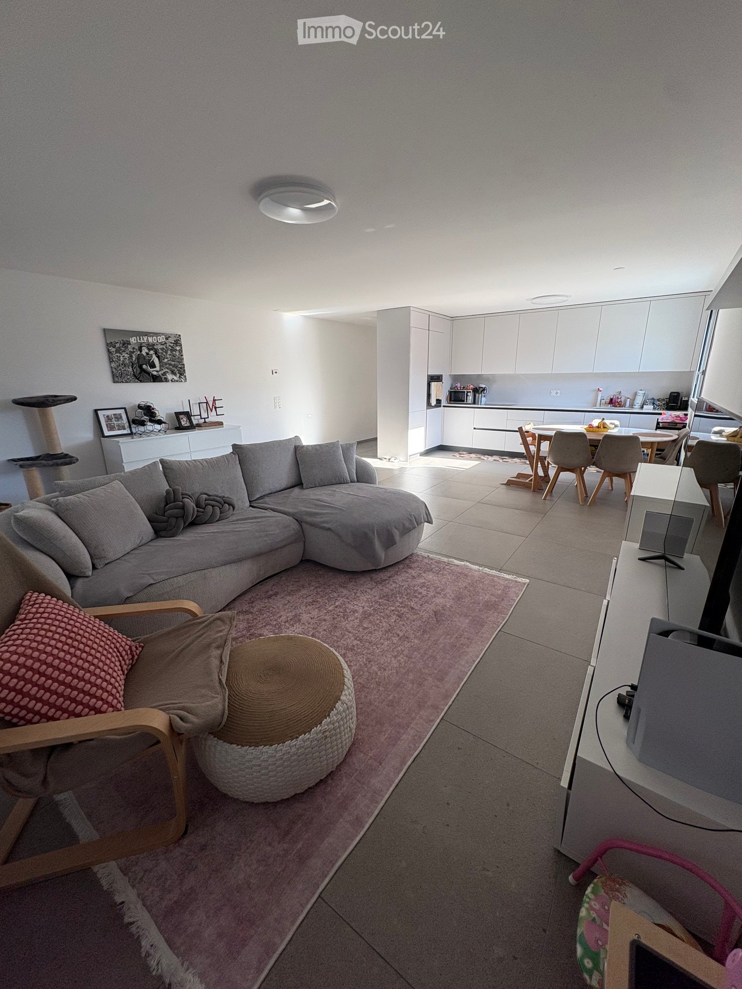 Modern living room with a grey sectional sofa, a round ottoman, a wooden armchair, a dining table with chairs, a cat tree, and a television. The space features a pink shag rug and white cabinets in the adjacent kitchen area.