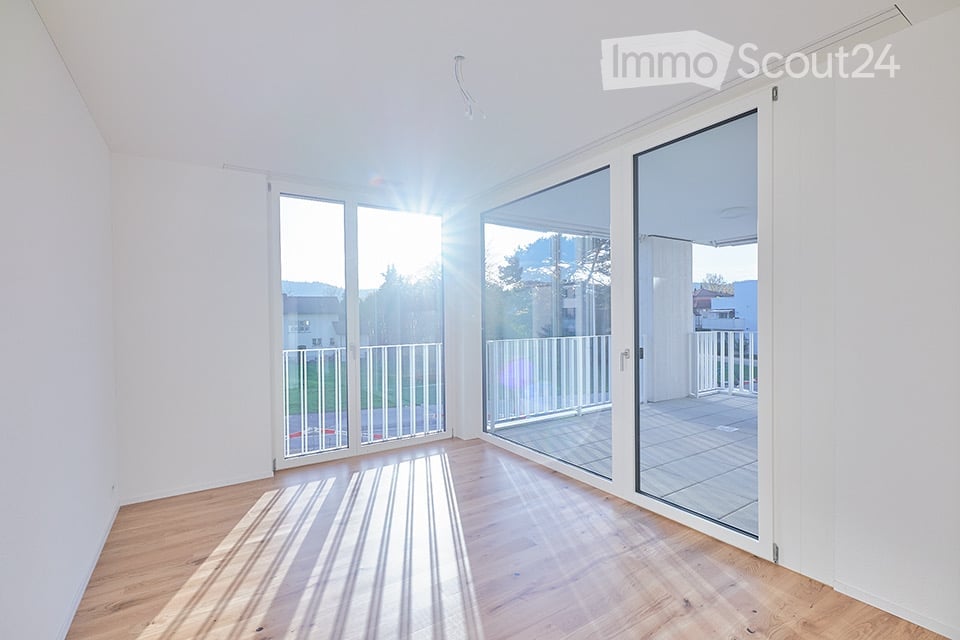 Bright, empty room with wooden flooring and large glass sliding doors leading to a balcony. Sunlight streams in, creating striped shadows on the floor.