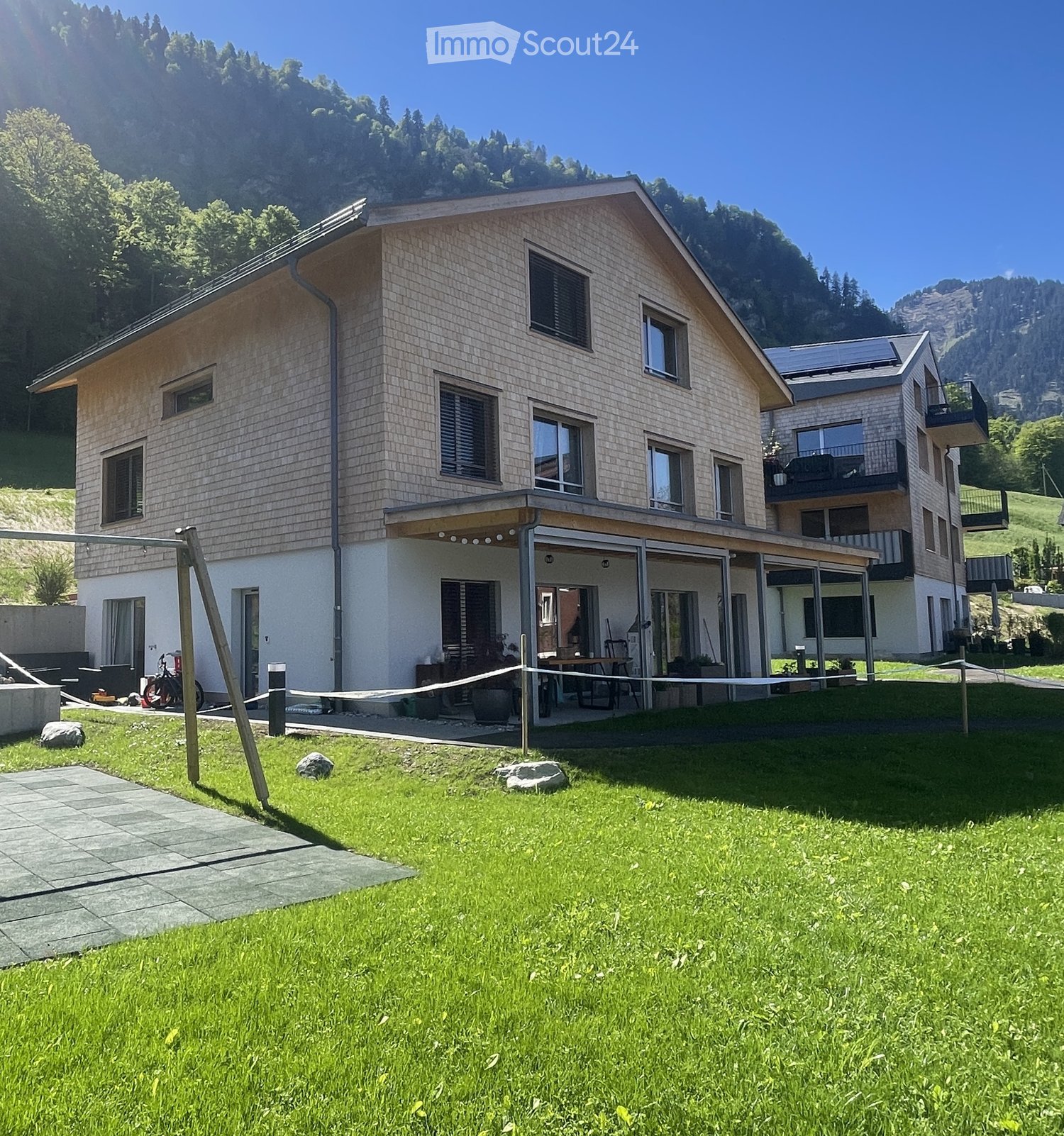 Two-story modern house with light-colored siding, large windows, and a spacious covered patio area. The house is surrounded by a well-maintained lawn and is situated in a scenic, mountainous area.