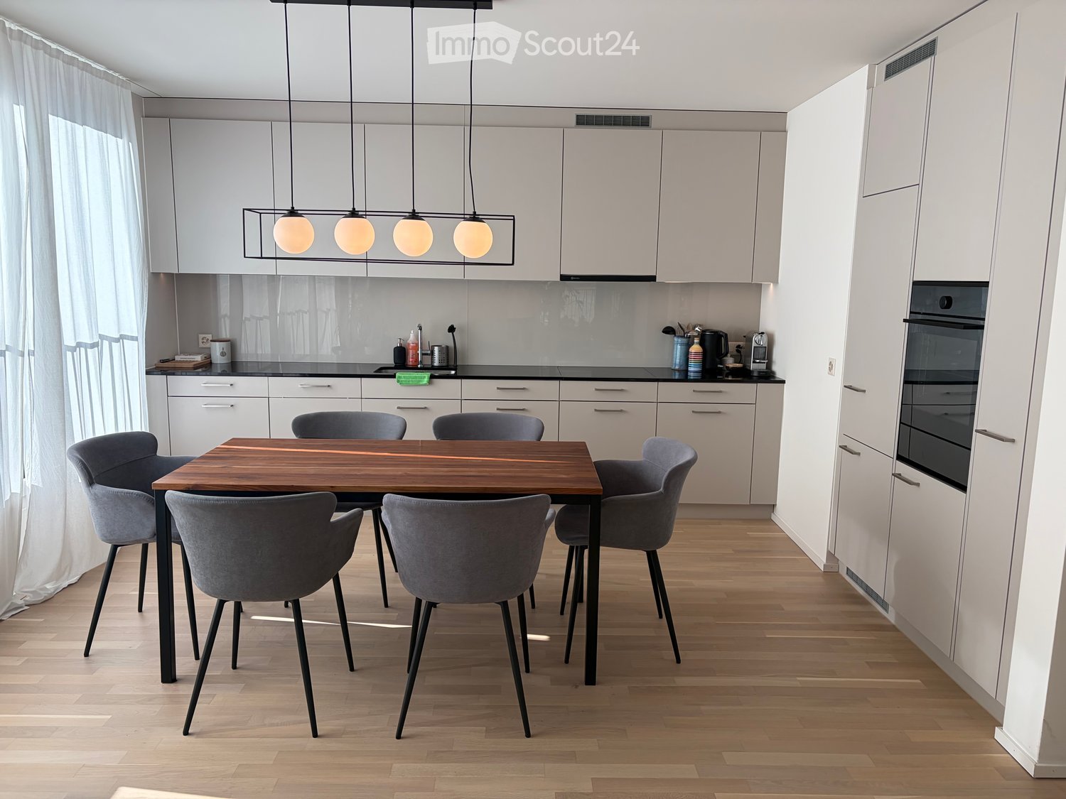 Modern kitchen with white cabinets, a wooden dining table surrounded by grey chairs, and various kitchen appliances including an oven and a coffee machine.