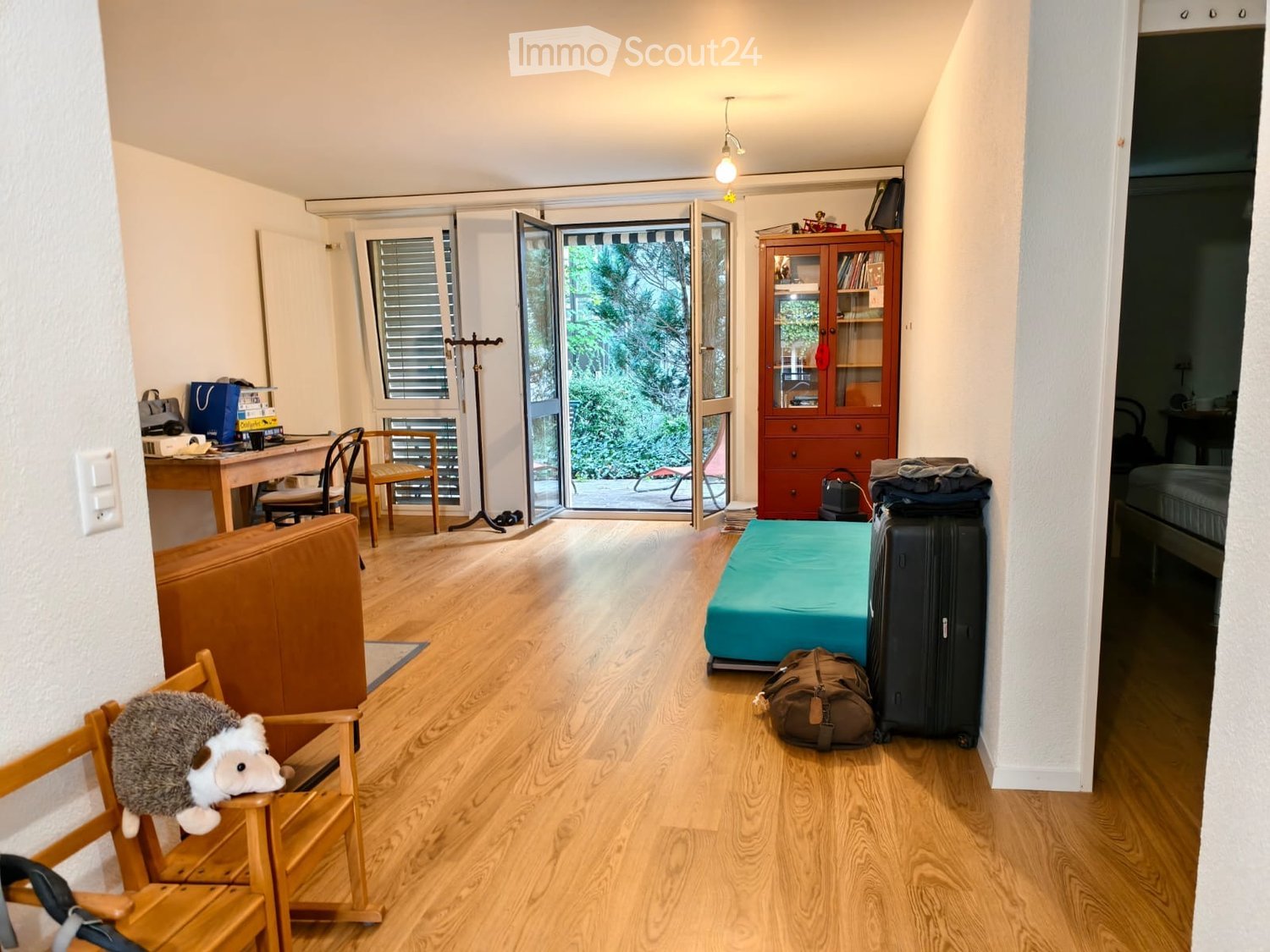 A furnished room with a sofa, chair, table, cabinet, stool, suitcase, backpack, and a toy on a chair. The room has wooden flooring and large windows with blinds.