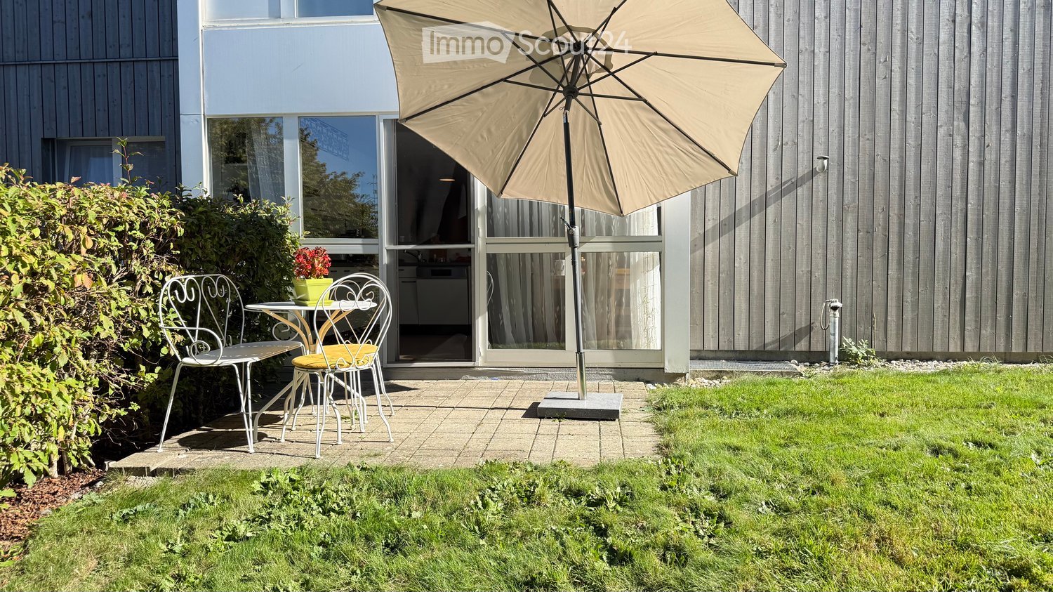 A small patio area with a table, two chairs, and an umbrella, adjacent to a modern house with large windows and a view of the interior.