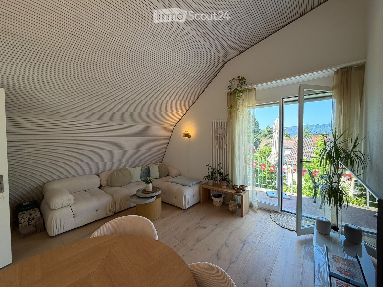 Bright living room with wooden flooring, a large window opening to a balcony, a cream-colored sofa, a round coffee table, a dining table with chairs, and several potted plants.