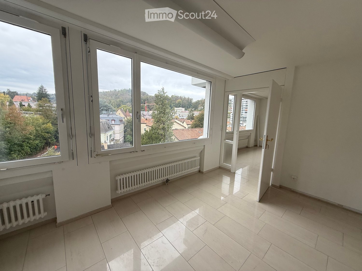 Empty room with tiled floor, large windows offering a view of trees and buildings, radiators along the walls, and an open doorway leading to another space.