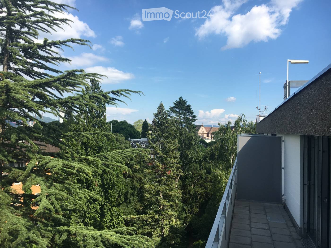 Balcony overlooking a lush, green garden with tall trees and a distant view of a building and rooftops under a clear blue sky.